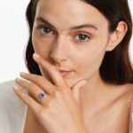 An 18K White Gold Oval Halo Engagement Rings With Diamonds and Sapphires ring featuring a 1.05 Carat Oval Blue Sapphire center stone and a 0.27 Carat Total Weight round diamond halo.