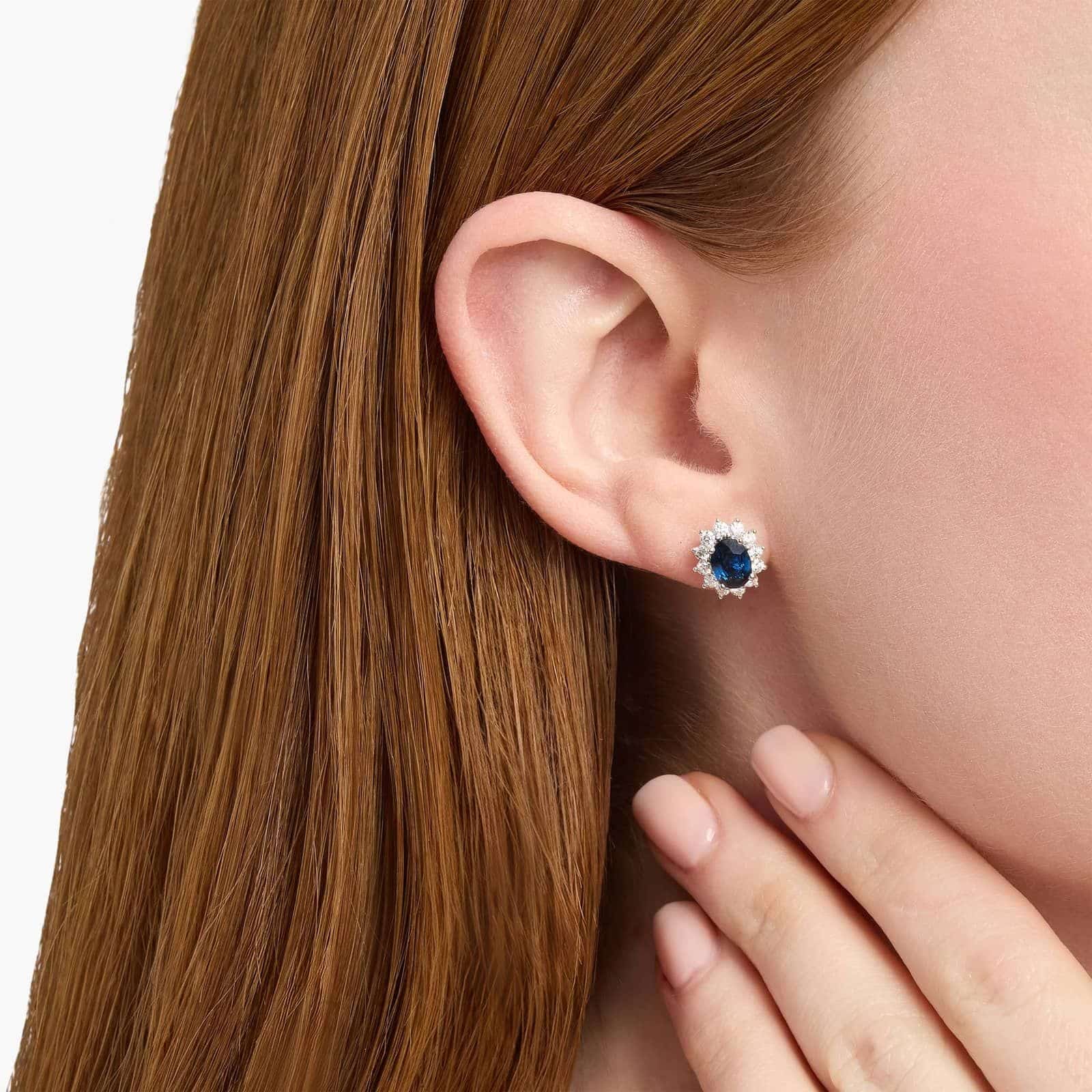 An 18K White Gold White Gold and Sapphire Earrings set featuring two 7x5 mm Oval Blue Sapphires surrounded by a 0.72 CTW Round Diamond Halo, secured with Push Backs.